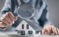 Person holding a magnifying glass over a model house, indicating inspection or investigation.